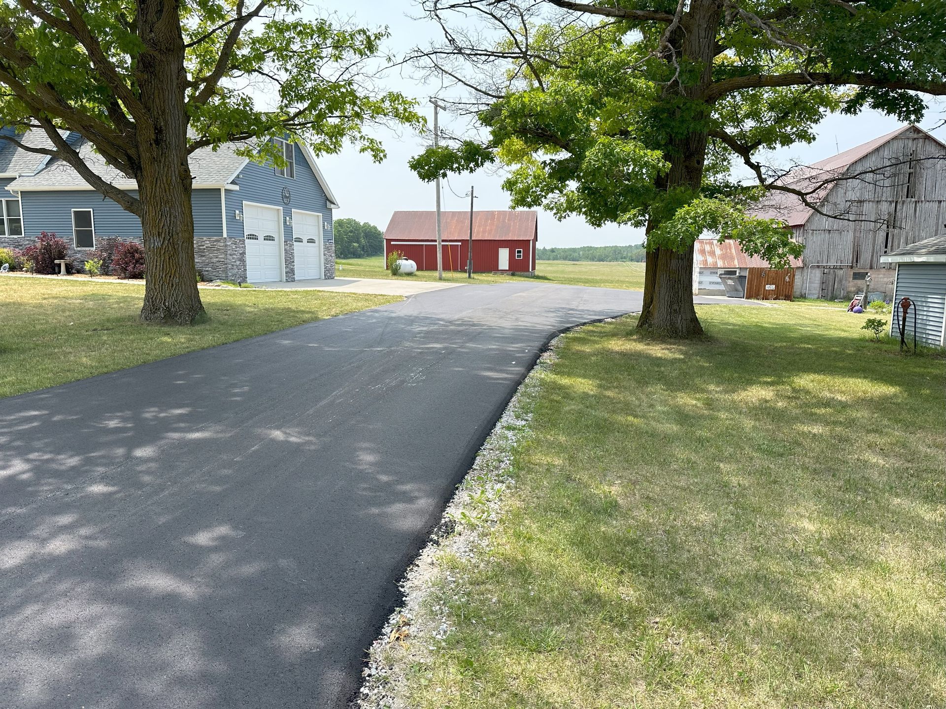 Paved driveway leading past trees and lawns to a red barn and gray outbuildings in a rural yard