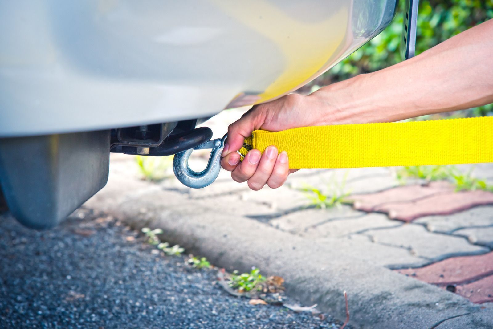 Person's hand attaching yellow tow strap to metal hook on car bumper outdoors.