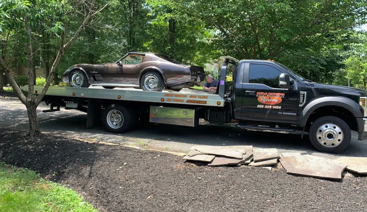 Tow truck carrying a vintage brown sports car along a wooded roadside