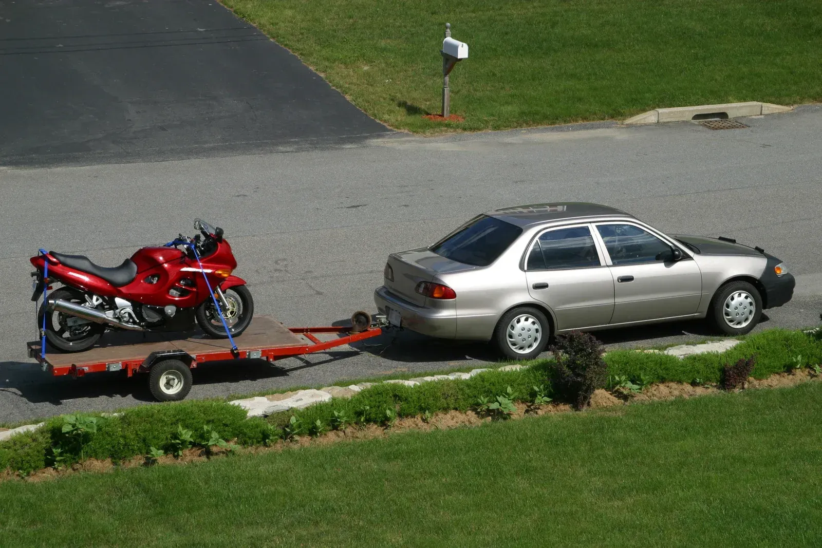 Red motorcycle on a trailer hitched to a silver car, parked on asphalt near grass.