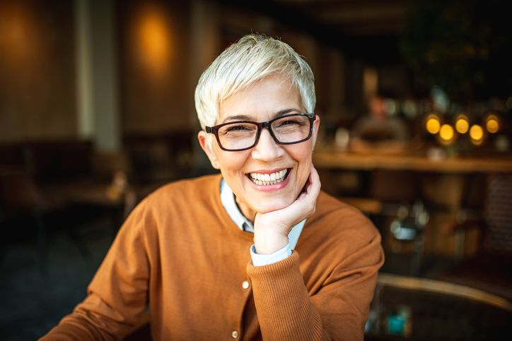 Woman with short, light-colored hair, wearing glasses and an orange sweater, smiling in a cafe.