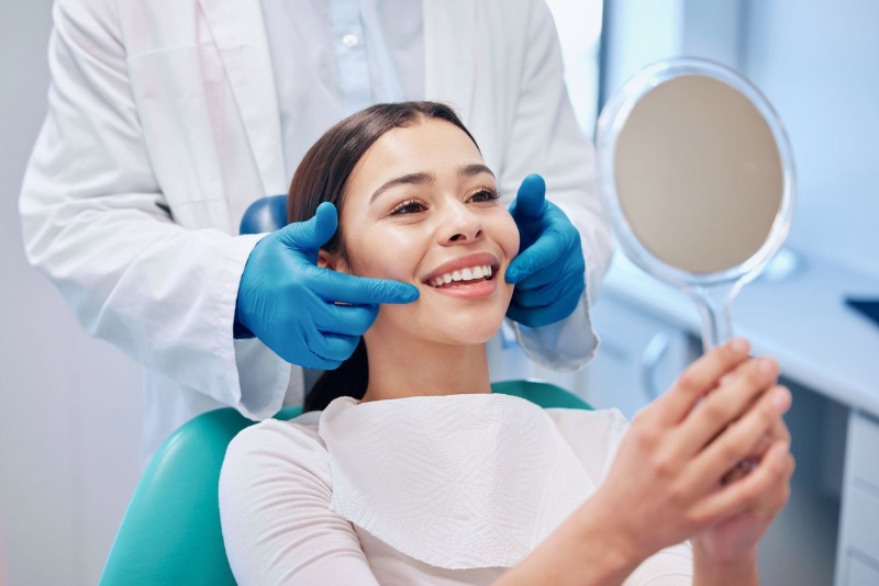 Woman in dentist chair, smiling while looking at her teeth in a mirror. Dentist examining her smile.