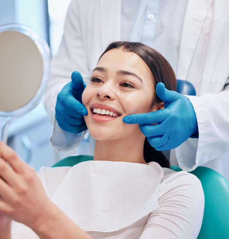 Woman smiles, looking in a mirror during a dental exam. Dentist in gloves examines her teeth in a clinic.