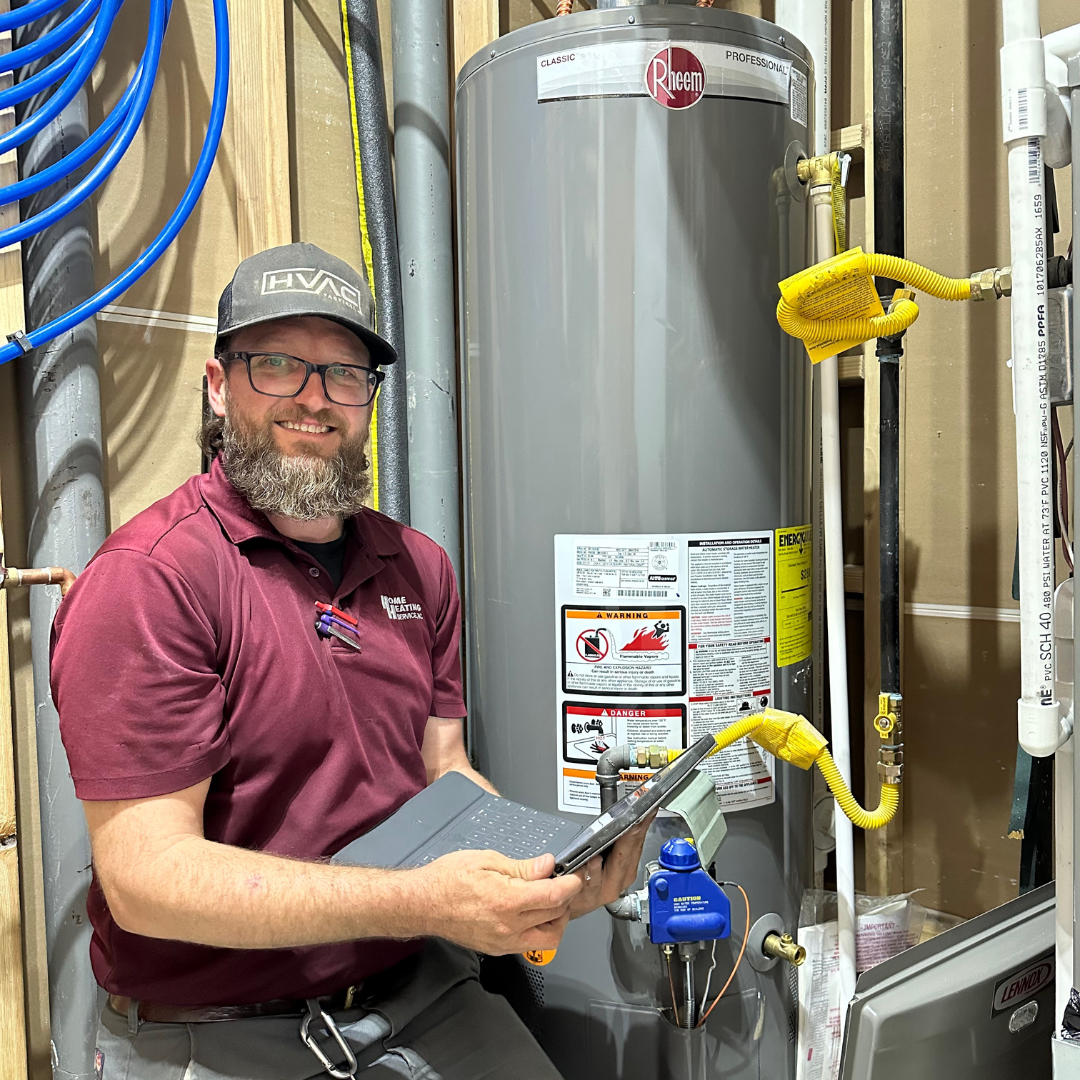 A smiling technician in a maroon polo and cap uses a tablet to inspect a large grey water heater in a mechanical room.