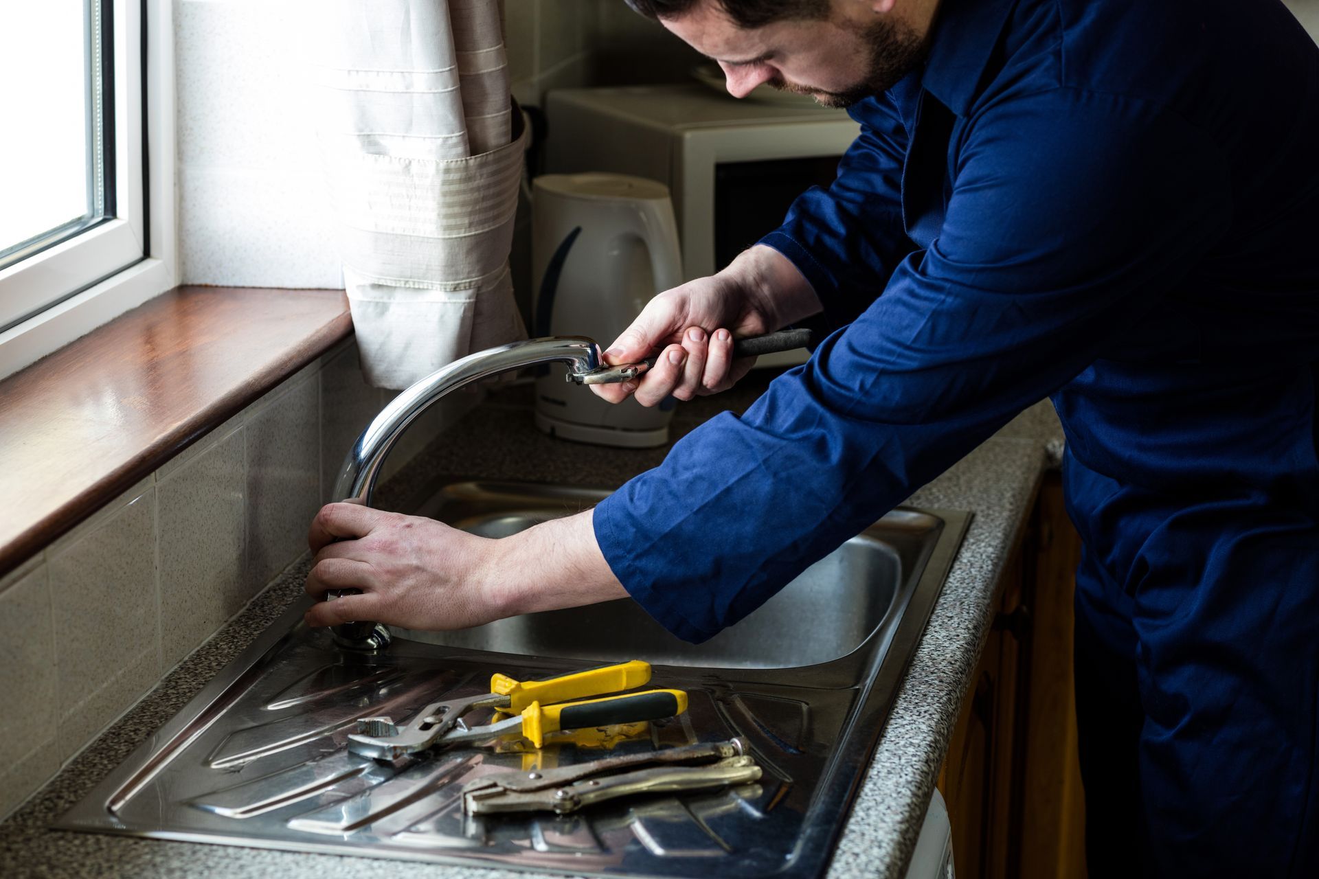 A plumber in blue coveralls tightens a kitchen faucet with pliers while plumbing tools sit nearby. A plumber in blue coveralls tightens a kitchen faucet with pliers while plumbing tools sit nearby.
