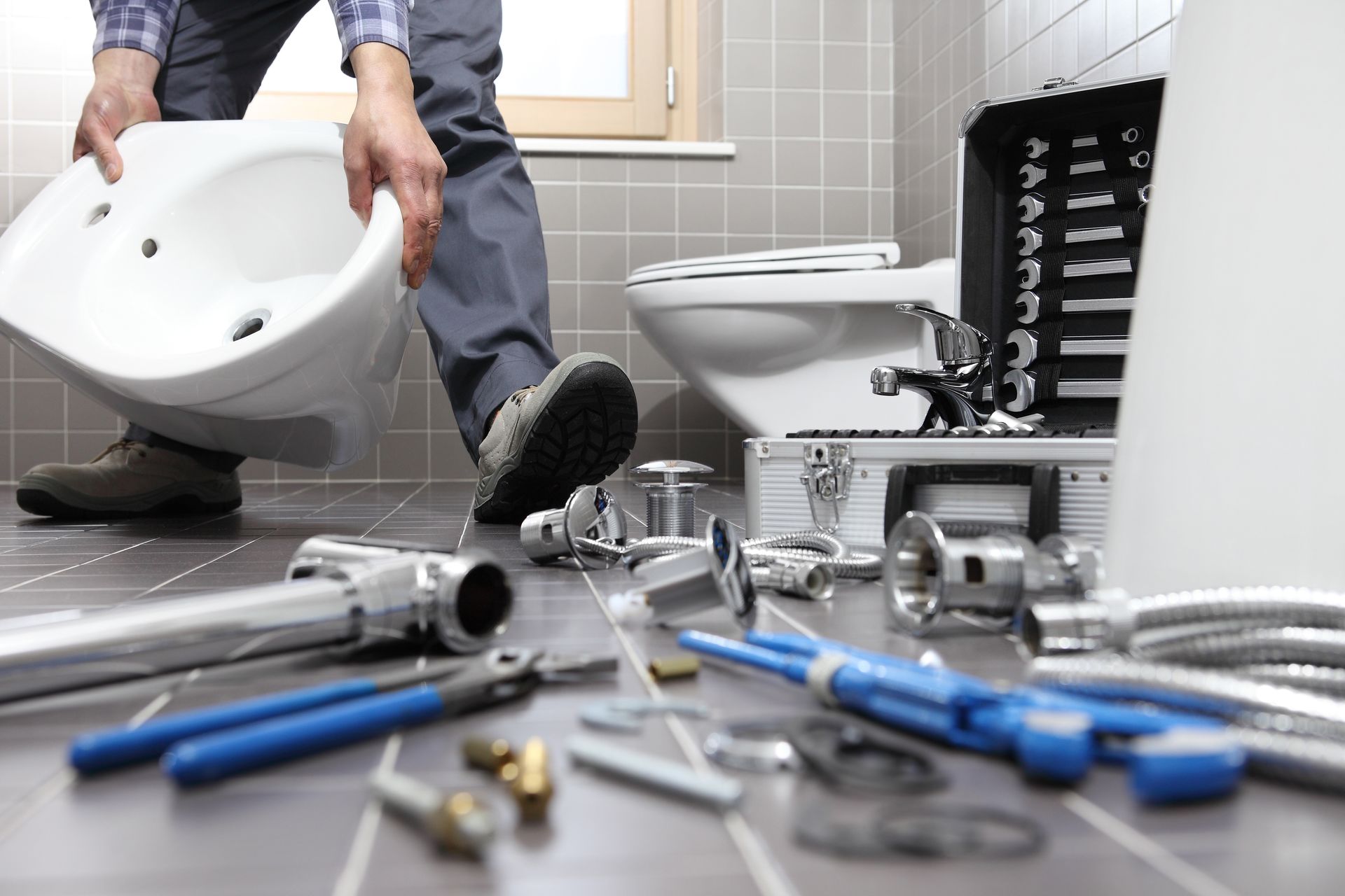 Plumber lifting a white sink in a tiled bathroom with plumbing tools and parts spread on the floor. Plumber lifting a white sink in a tiled bathroom with plumbing tools and parts spread on the floor.
