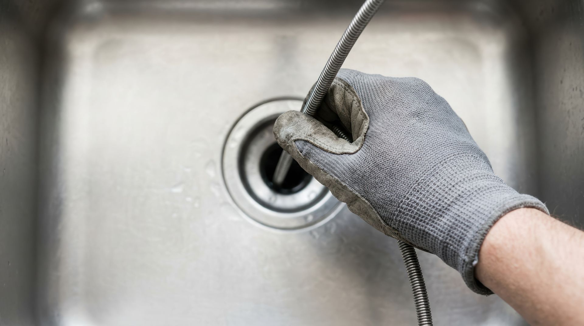 Gloved hand feeding a coiled metal drain snake into a stainless-steel sink during drain cleaning. Gloved hand feeding a coiled metal drain snake into a stainless-steel sink during drain cleaning.