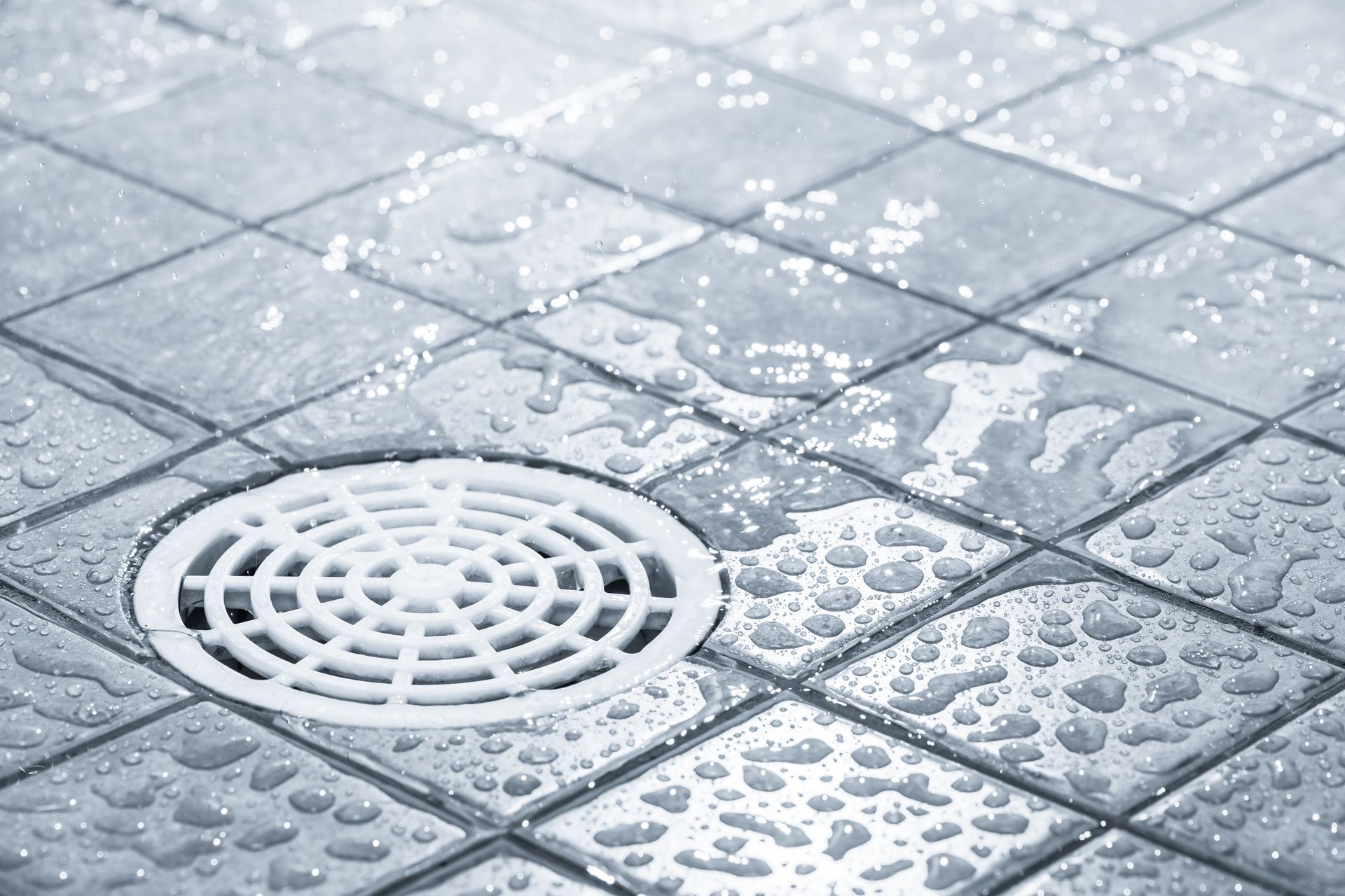 Wet tiled floor with water droplets and a round drain grate in a bathroom. Wet tiled floor with water droplets and a round drain grate in a bathroom.