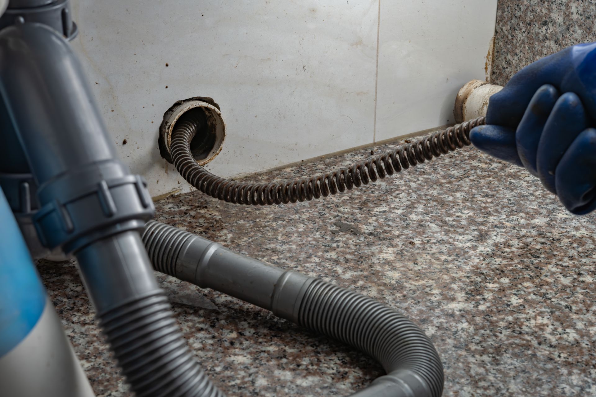 Gloved plumber feeding a drain cleaning cable into a wall pipe opening next to gray flexible hoses.
