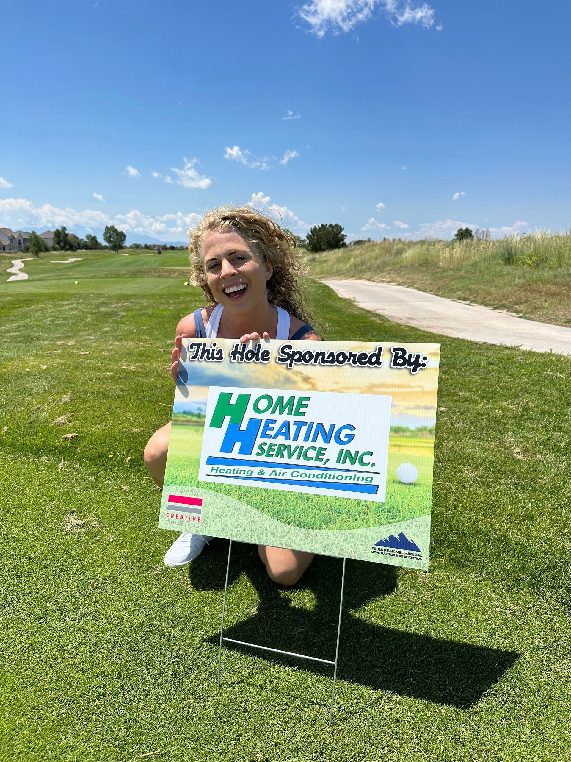 A person kneeling on a golf course holding a sponsorship sign for Home Heating Service, Inc. under a bright blue sky.