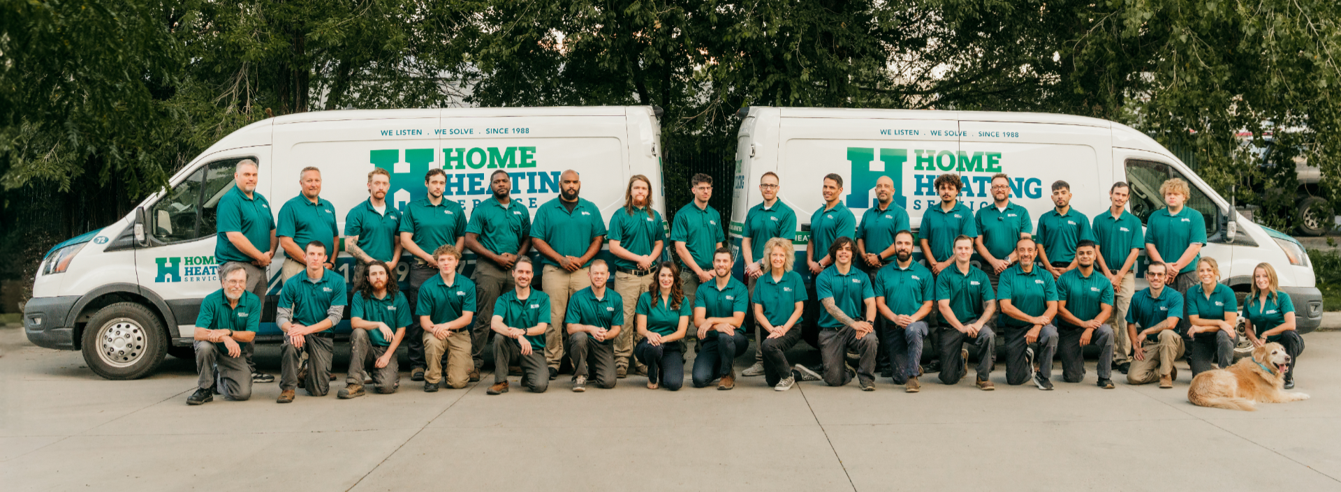 A large group of people in branded teal shirts posing with a dog in front of two Home Heating Service vans.