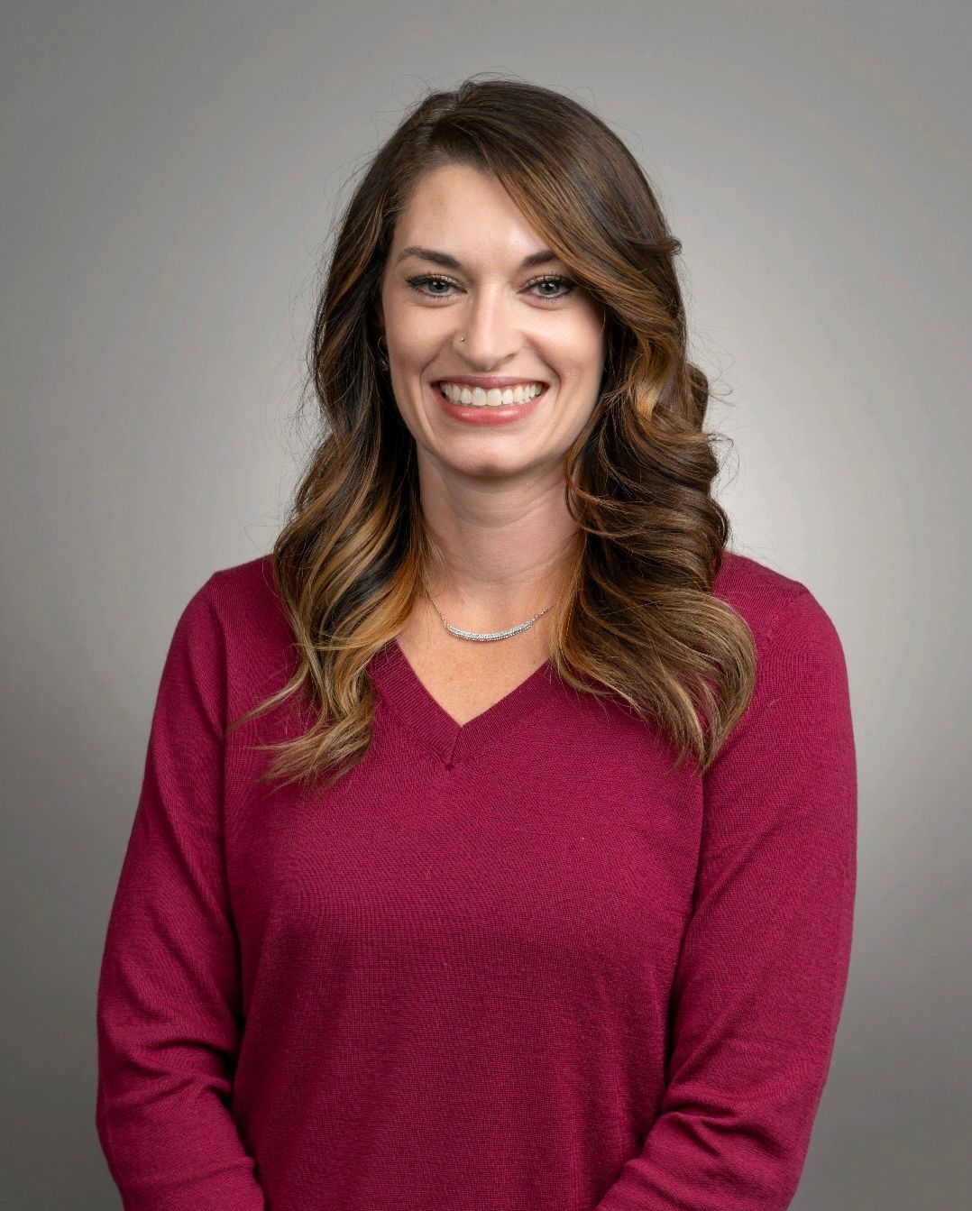 A smiling person with shoulder-length wavy brown hair wearing a long-sleeved magenta sweater against a gray background.