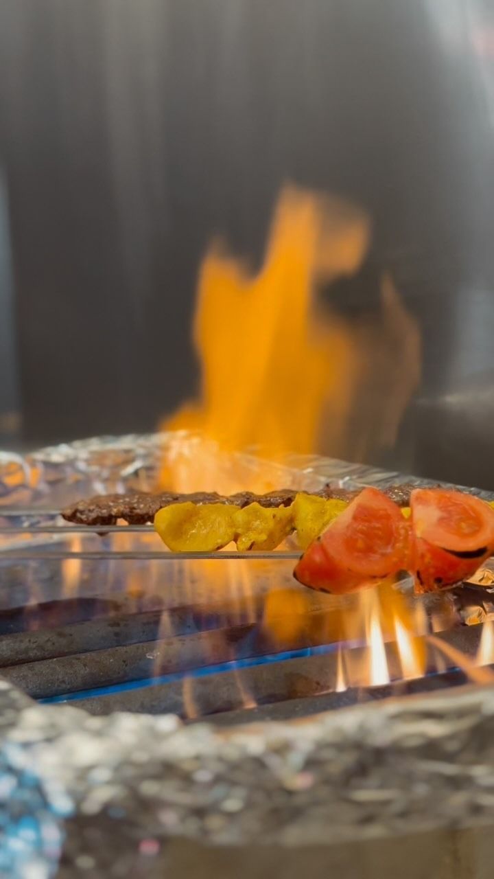 Carne allo spiedo, verdure gialle e fette di pomodoro grigliate su fiamme libere.