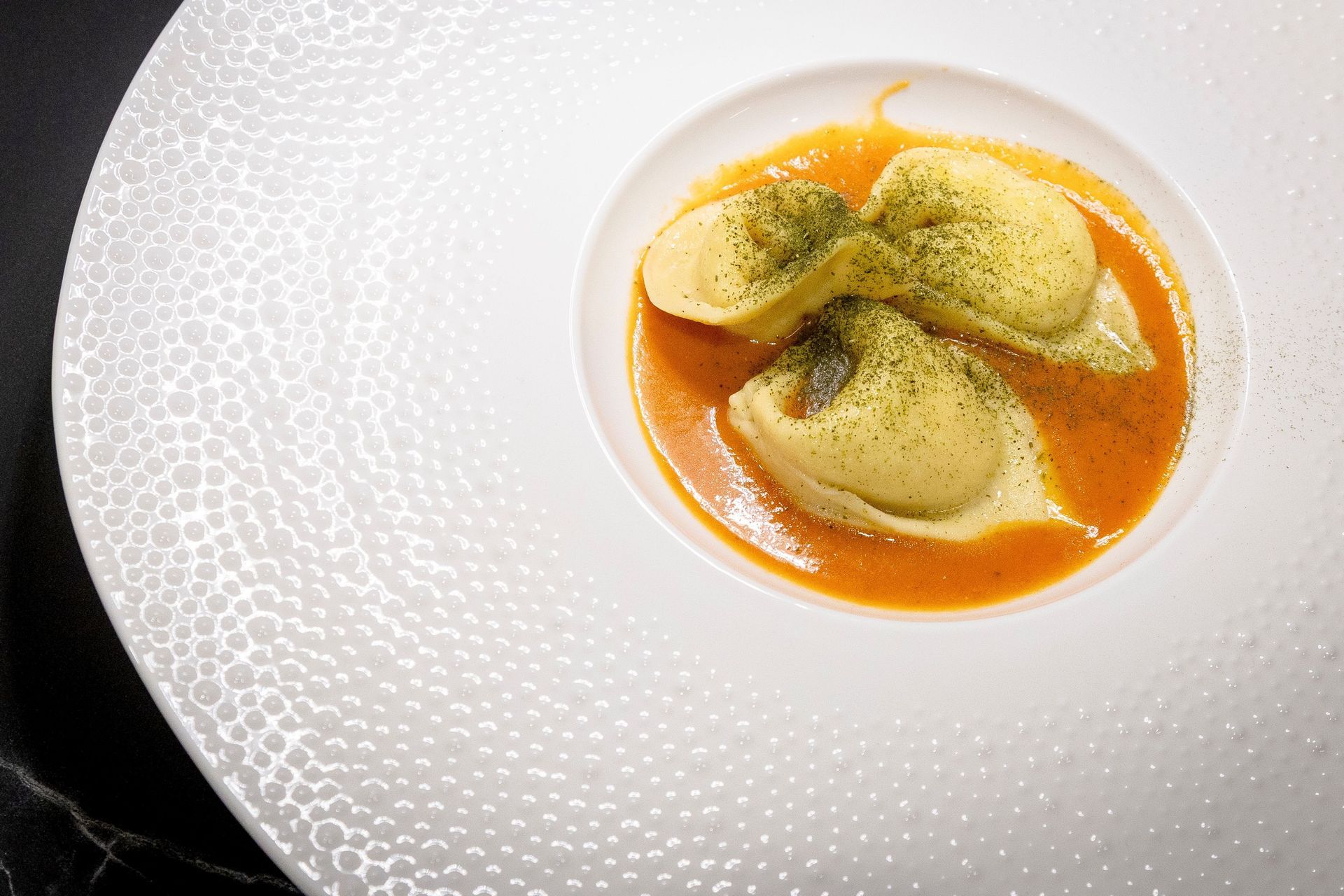 Ravioli in salsa all'arancia, guarniti con briciole di erbe aromatiche, su un piatto bianco punteggiato.