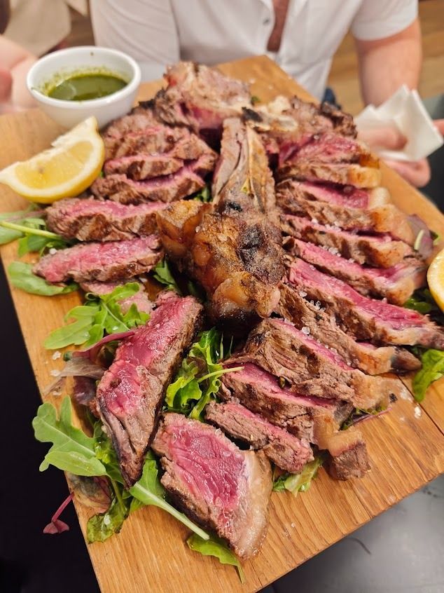 Piatto di bistecca grigliata a fette con erbe aromatiche, spicchi di limone e salsa verde su un tagliere di legno.