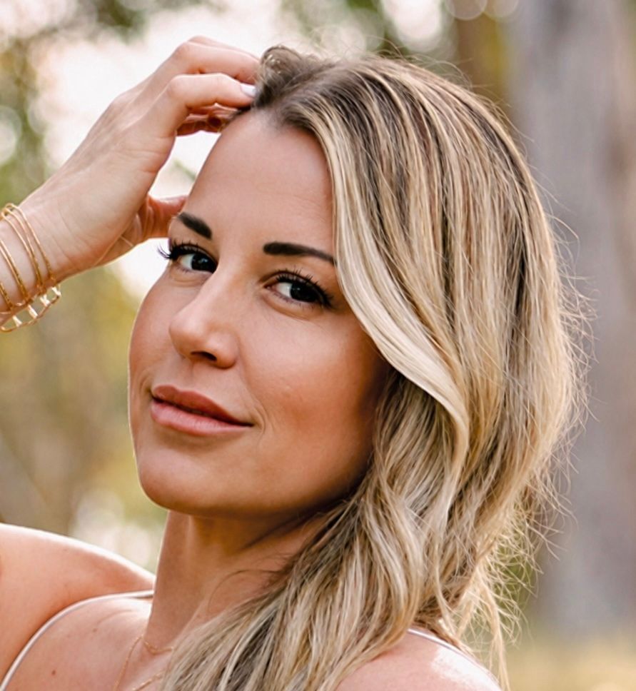 Blonde woman in a white top touching her hair outdoors, looking at the camera.