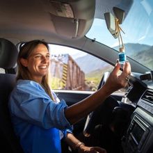 Una mujer sonríe dentro de un coche mientras sostiene en alto un vial azul, con las montañas visibles.