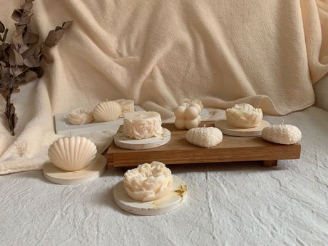 Conchas blancas variadas y jabones florales sobre una bandeja de madera, encima de una suave tela beige.