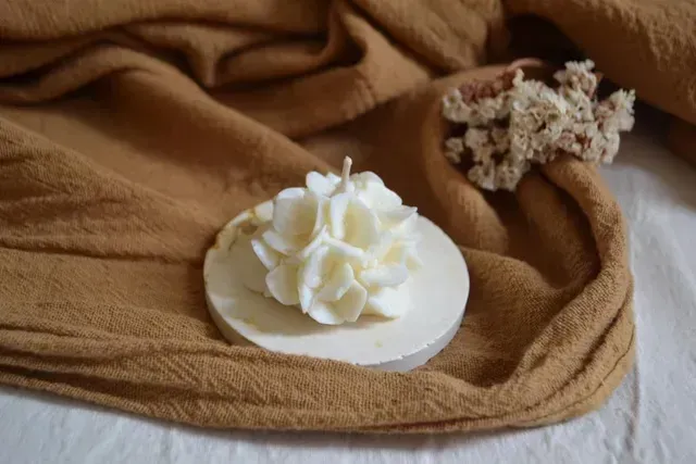 Vela blanca con forma de flor sobre una tela beige, junto a flores secas y una manta tejida color tostado