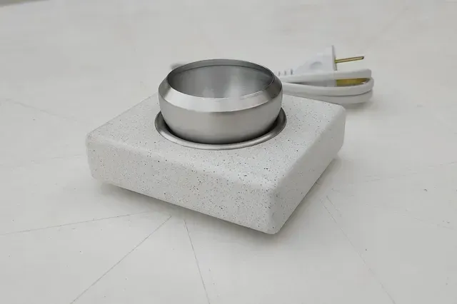 Pequeño dispositivo cuadrado blanco con un vaso metálico y un cable de alimentación sobre una superficie clara.