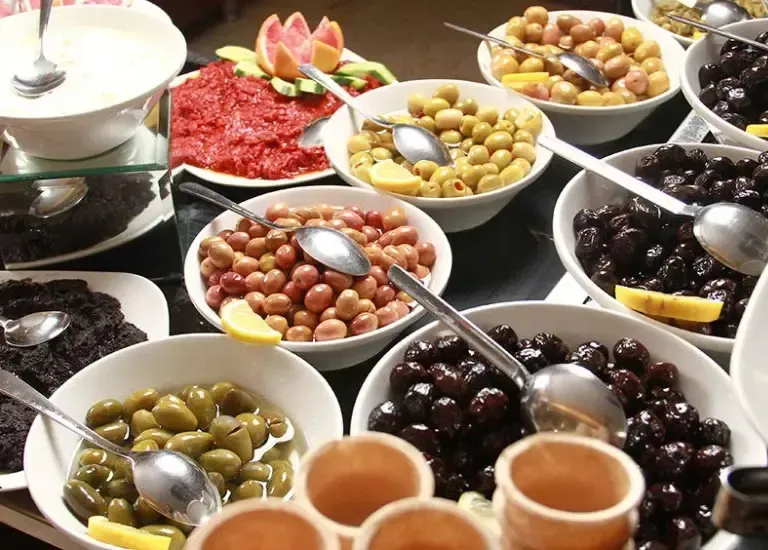 A buffet table spread featuring various bowls of olives, spreads, and fruit with serving spoons in a dining setting.