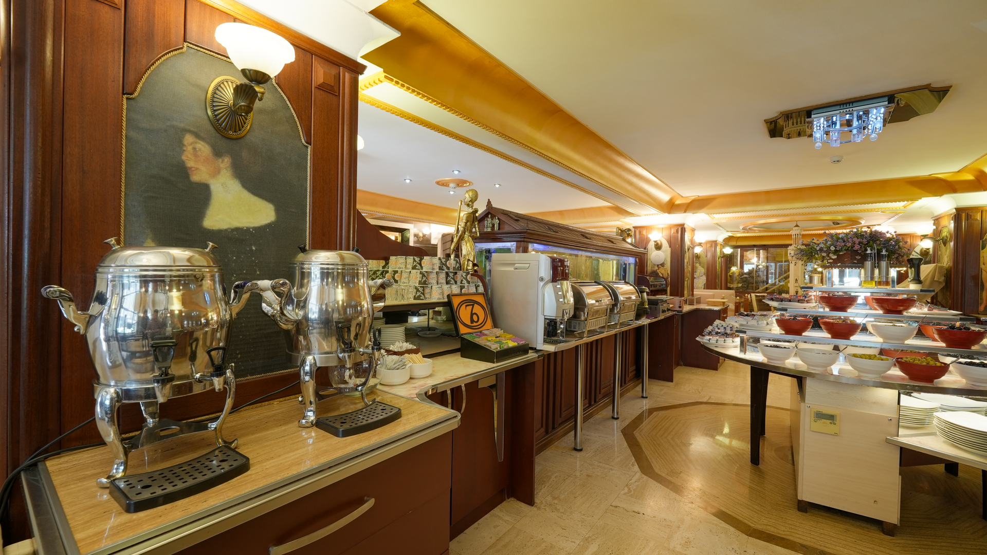 A buffet breakfast area featuring metallic hot beverage urns, food warming stations, and tables with dishes in a dining room.