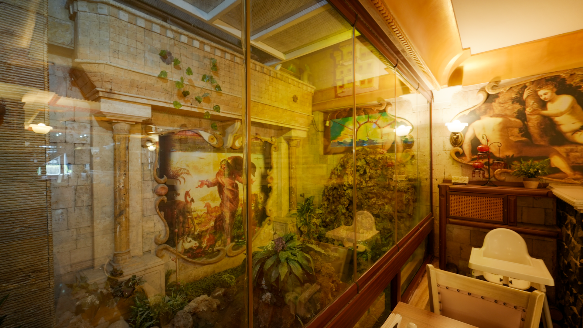 An indoor glass-enclosed display featuring a classical architectural backdrop, a mural of figures, and lush greenery.