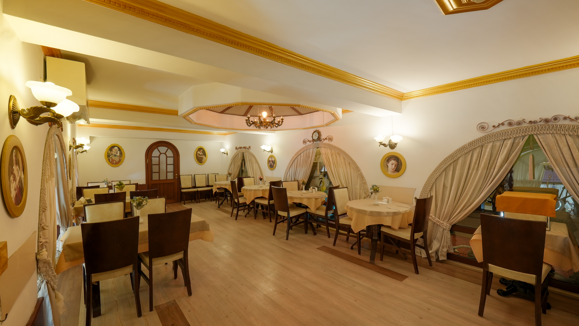 An elegant, well-lit restaurant dining room with cream-colored walls, gold ceiling trim, draped curtains, and wood chairs.