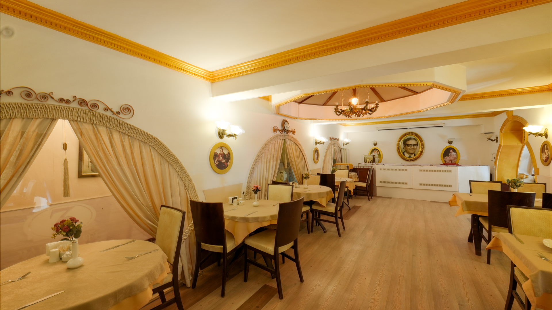 A dining room featuring several tables with beige tablecloths, dark chairs, and decorative curtain arches in warm tones.
