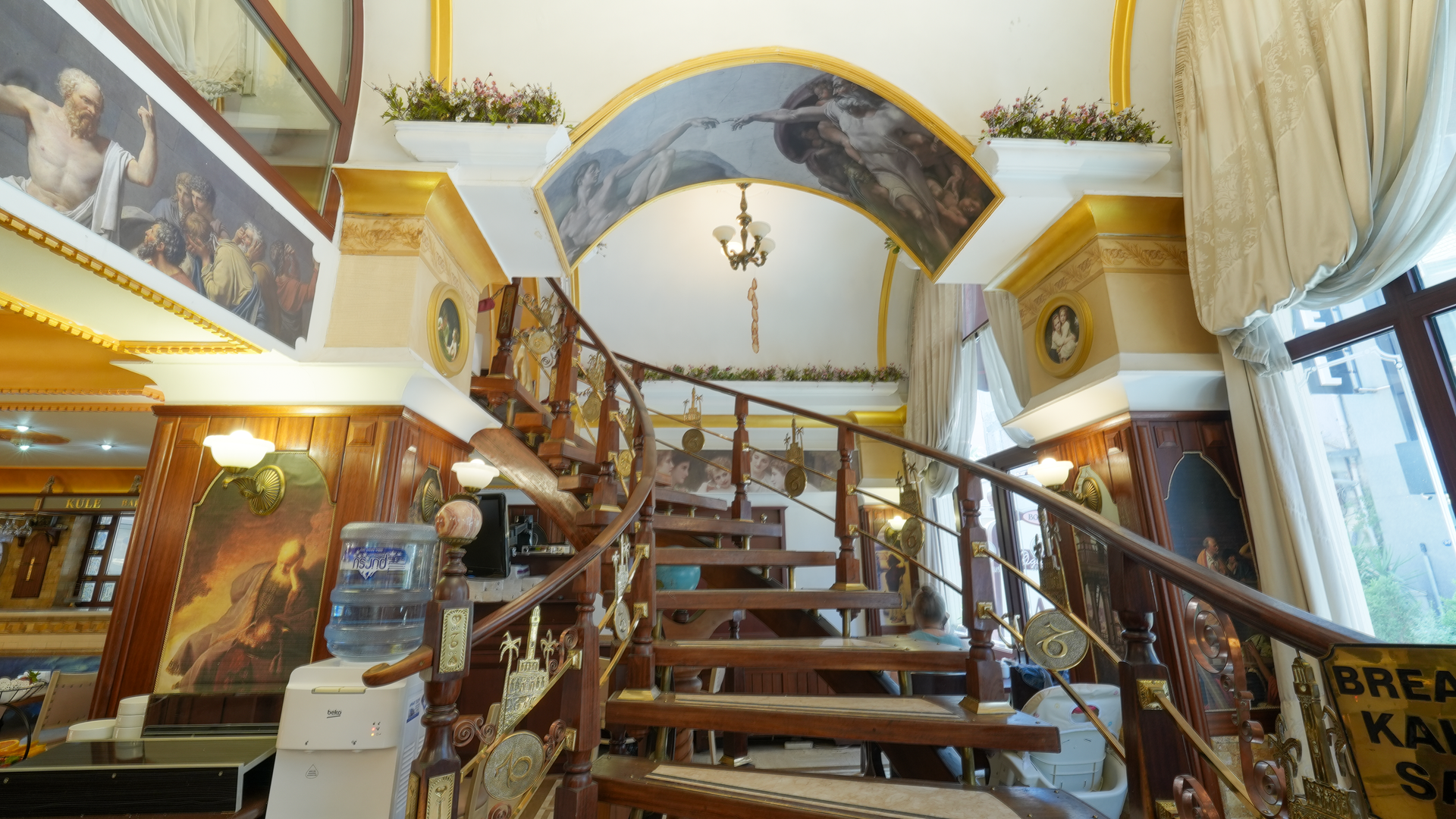 A decorative wooden spiral staircase inside a grand lobby, framed by a painted archway and ornate wall murals.