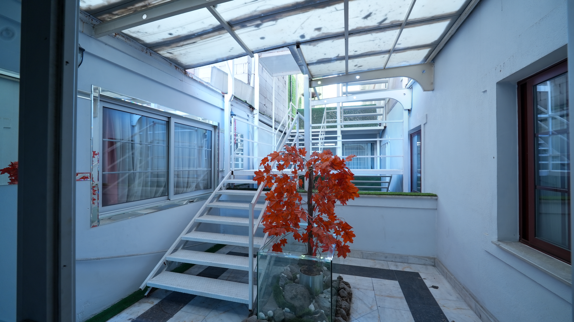 A white metal staircase sits in a bright, enclosed courtyard with a translucent roof and an orange artificial tree.