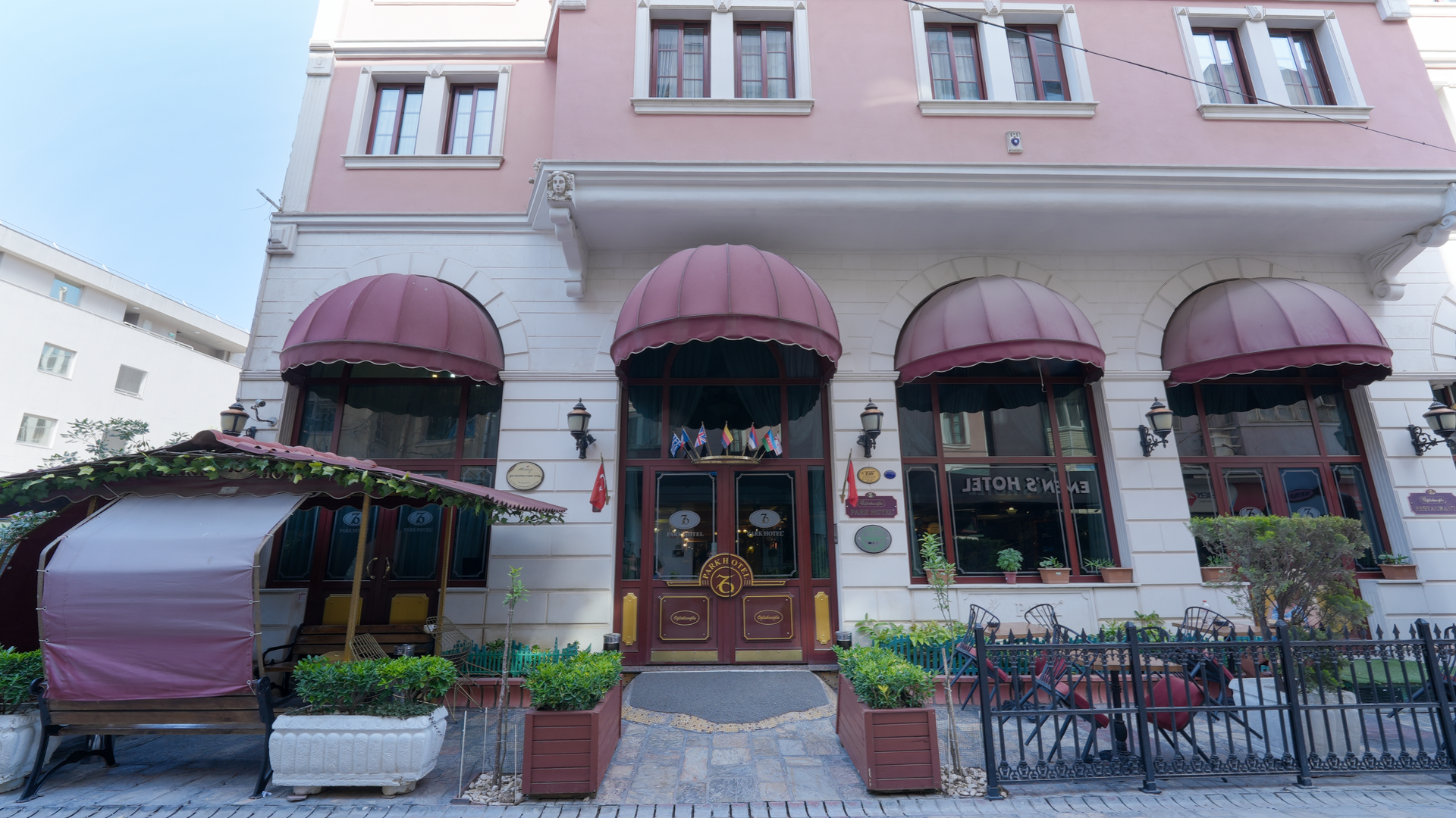  İzmir Park Hotel. The facade of a pink and white building with arched windows, maroon awnings, and an outdoor seating area.