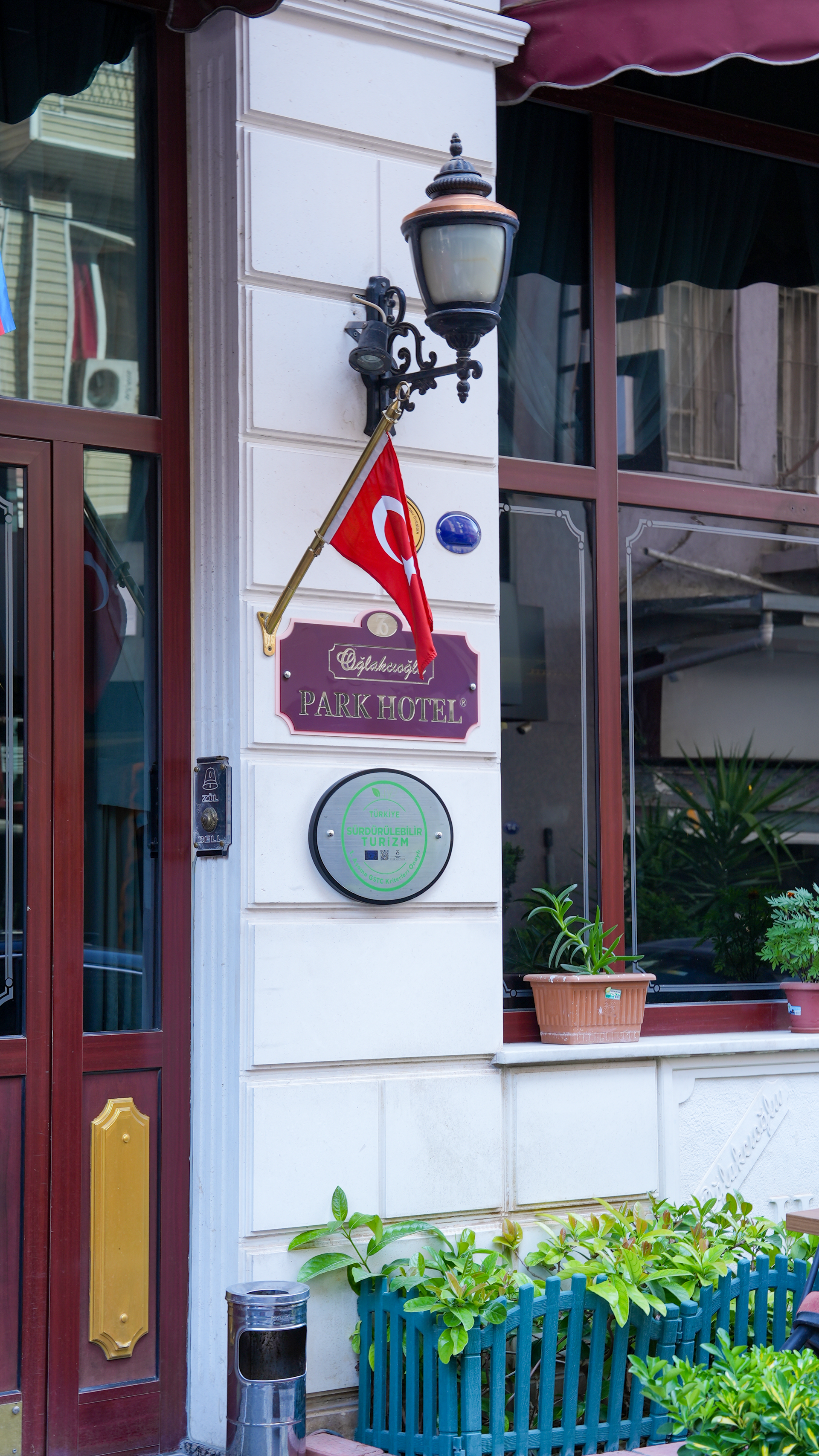A Turkish flag hangs under a street lamp on the white exterior wall of a building near a door and a window. Oğlakçıoğlu İzmir