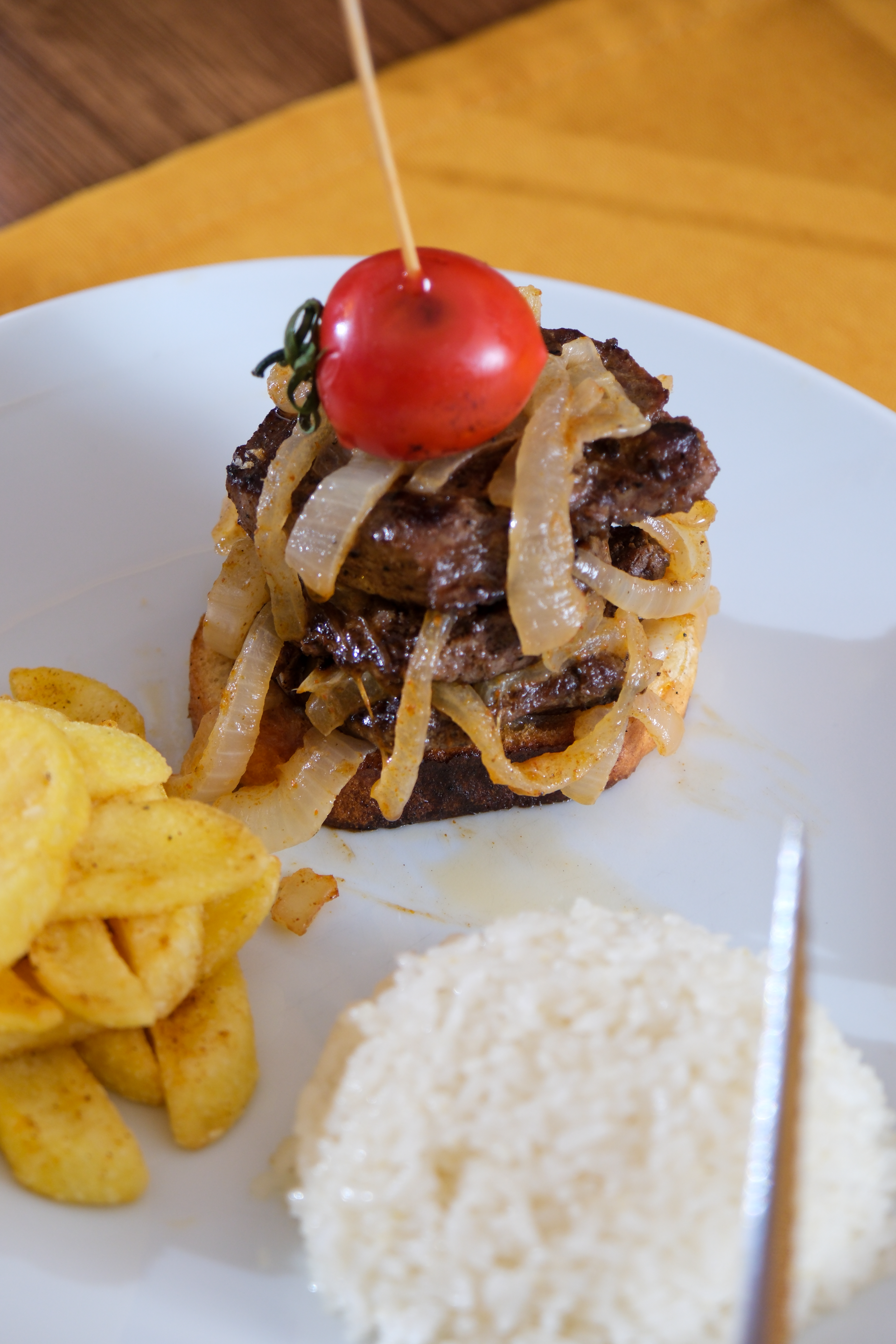 A steak sandwich topped with sautéed onions and a cherry tomato, served with a scoop of white rice and potato wedges.