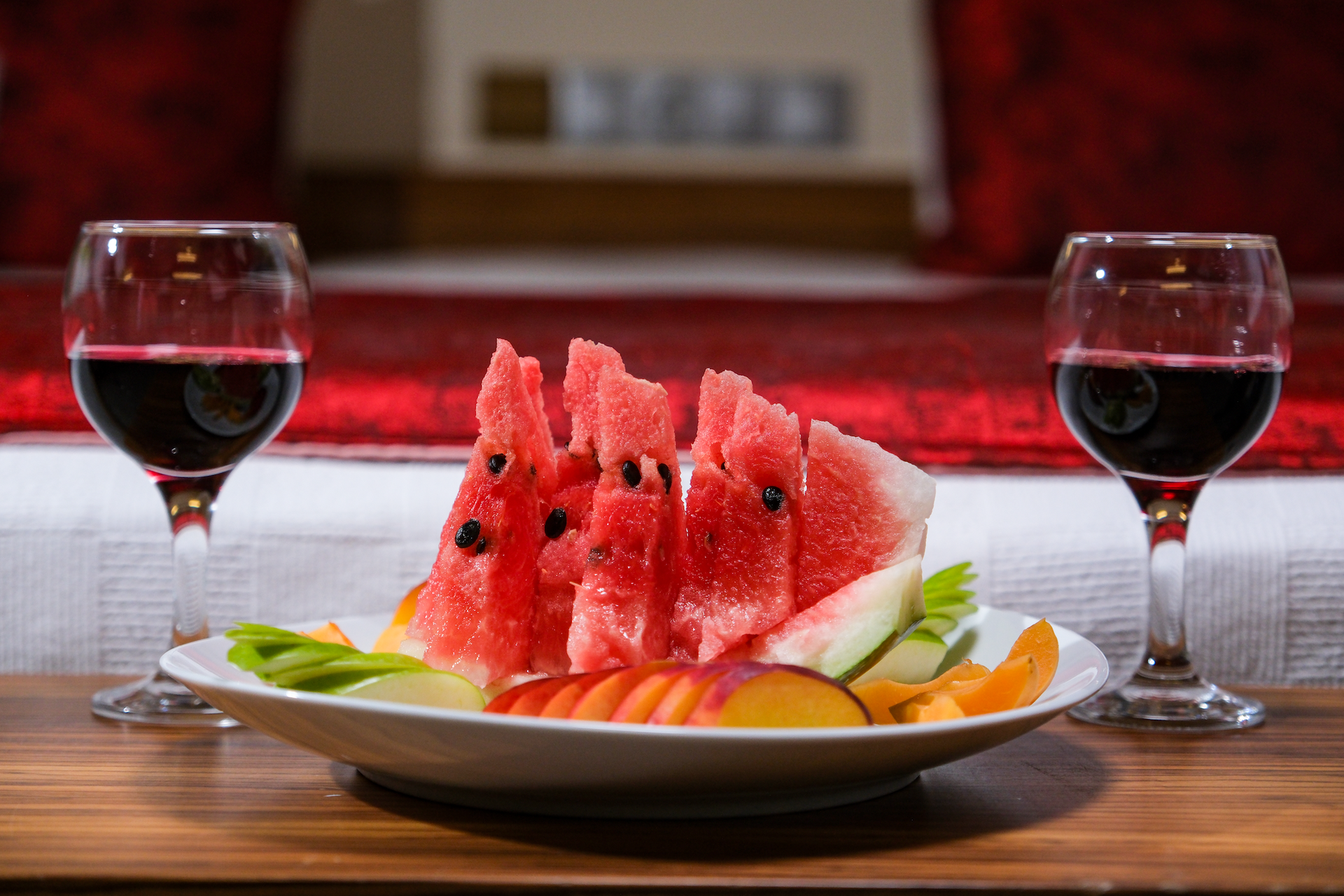 A plate of fresh sliced watermelon, kiwi, and nectarines sits between two glasses of red wine on a wooden table.