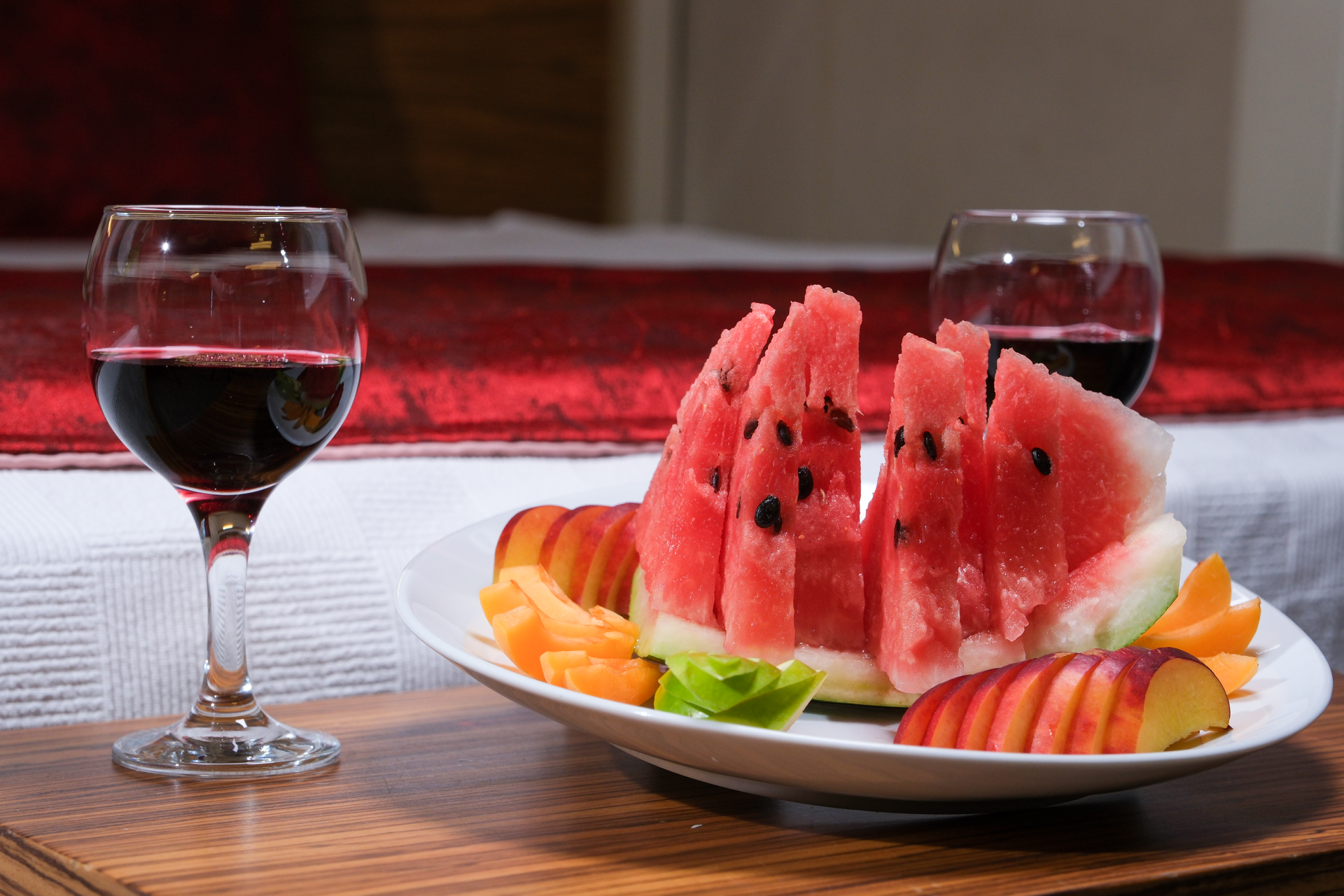 A plate of watermelon slices and nectarines sits on a table next to two glasses of red wine in front of a made bed.