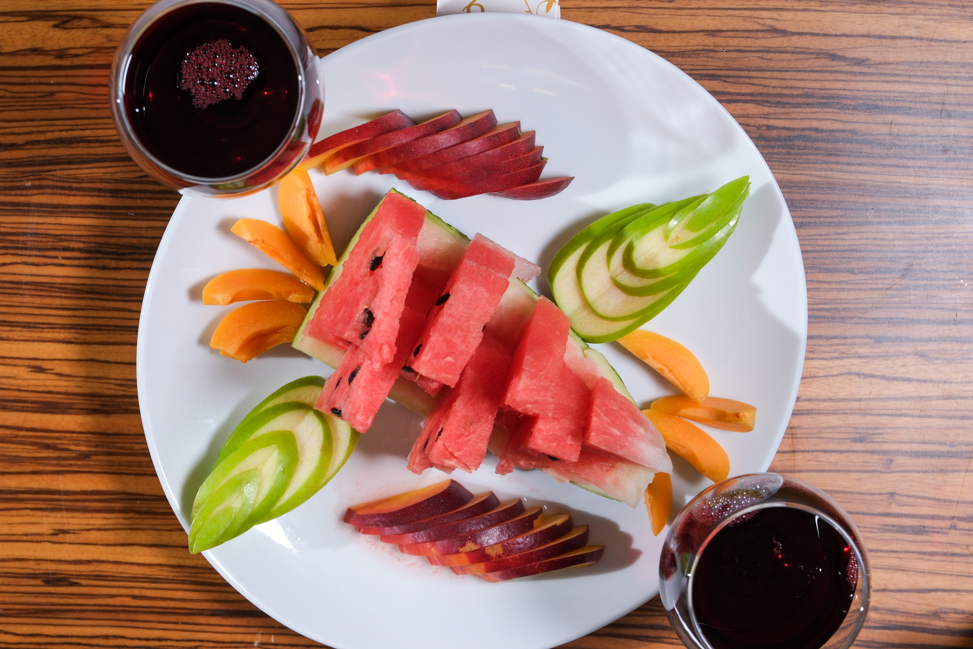 A white plate features sliced watermelon, apple fans, and orange segments, flanked by two glasses of dark red wine.