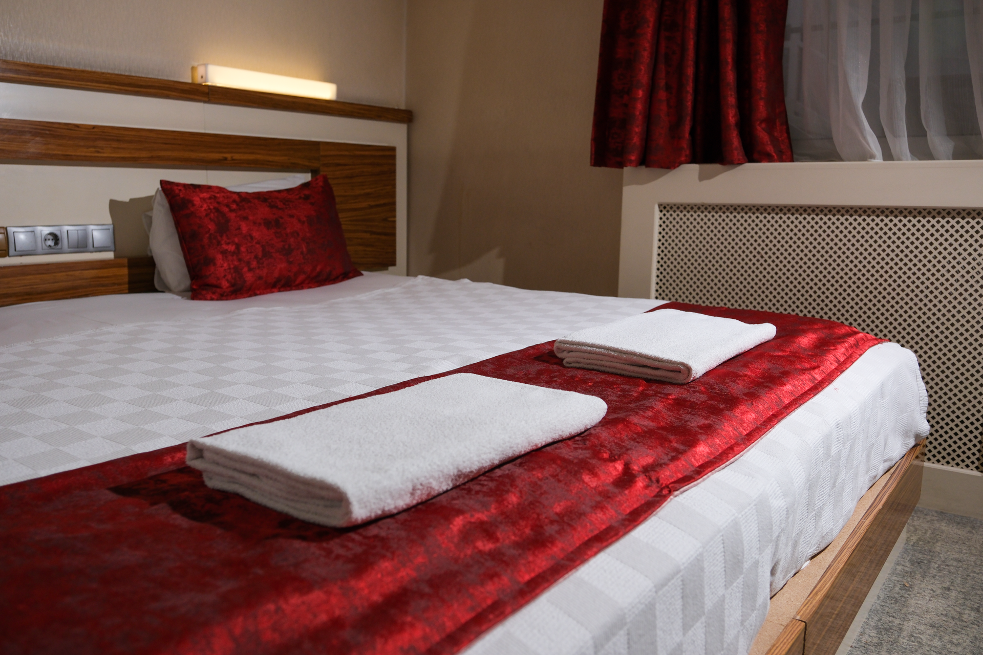 A made hotel bed with white linens, a red runner, two folded white towels, and red accent pillows against a wooden headboard.