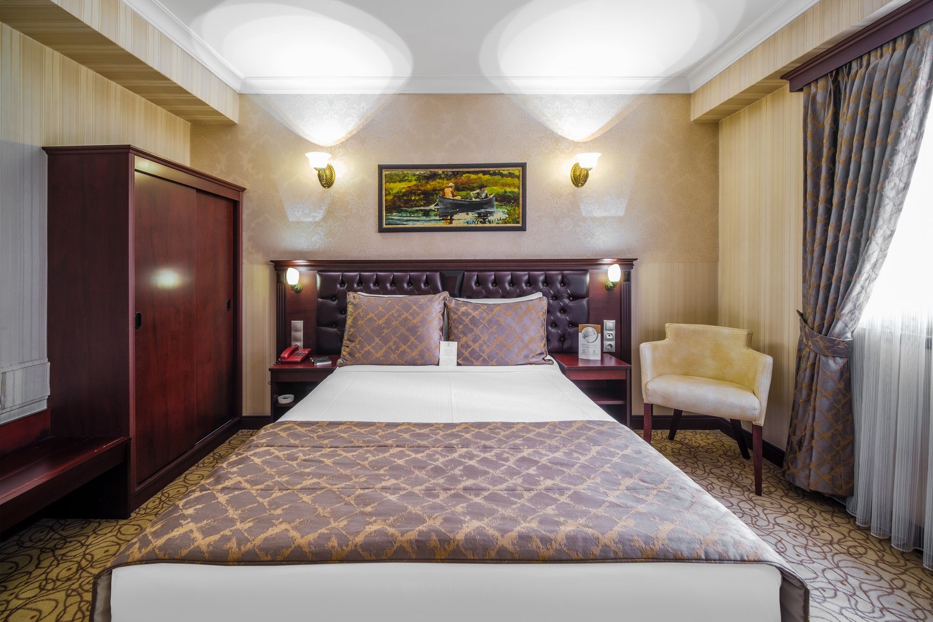 İzmir Park Hotel's tidy room with a queen bed, dark wood wardrobe, patterned runner, wall art, and an armchair by a window.