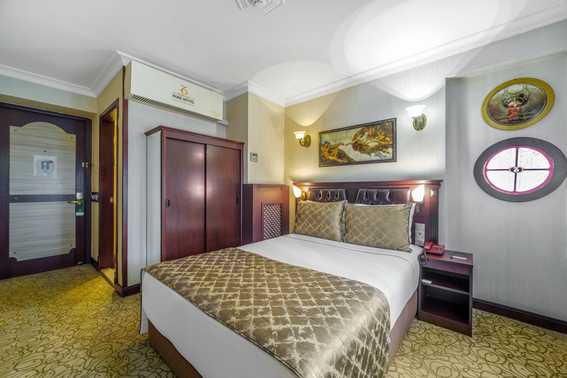 A compact hotel room with a neatly made bed, a wooden wardrobe, wall art, a circular window, and patterned beige carpet.