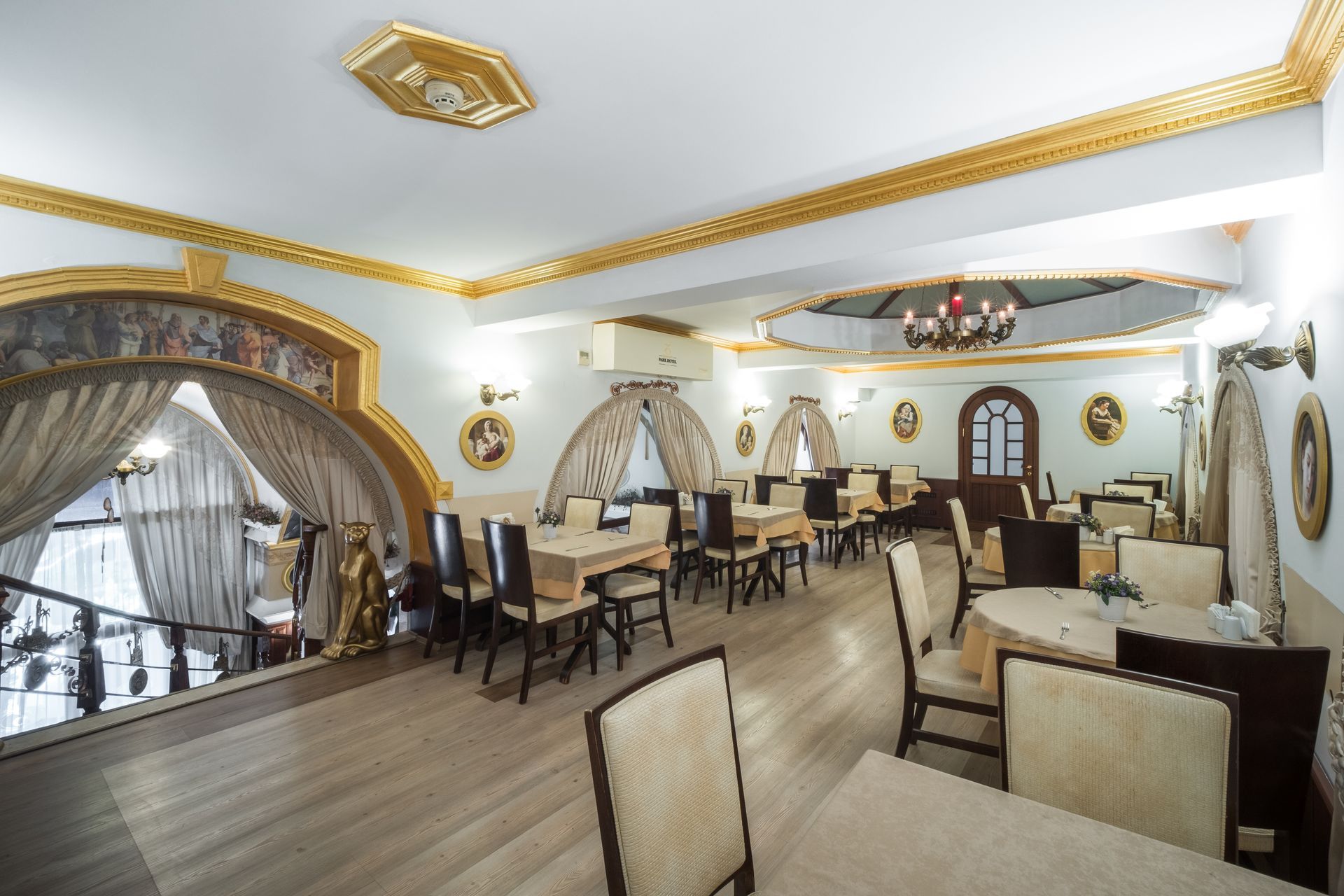 An elegant, light-filled dining room with beige chairs, round tables, arched architectural features, and gold trim.