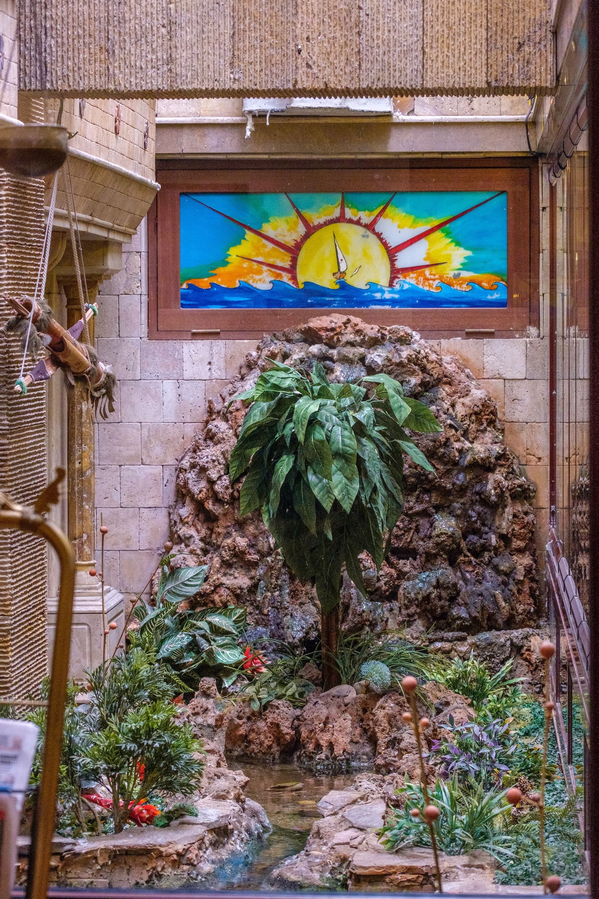 A stone indoor grotto with a lush green plant, a small pool of water, and a sun-themed stained glass window above.