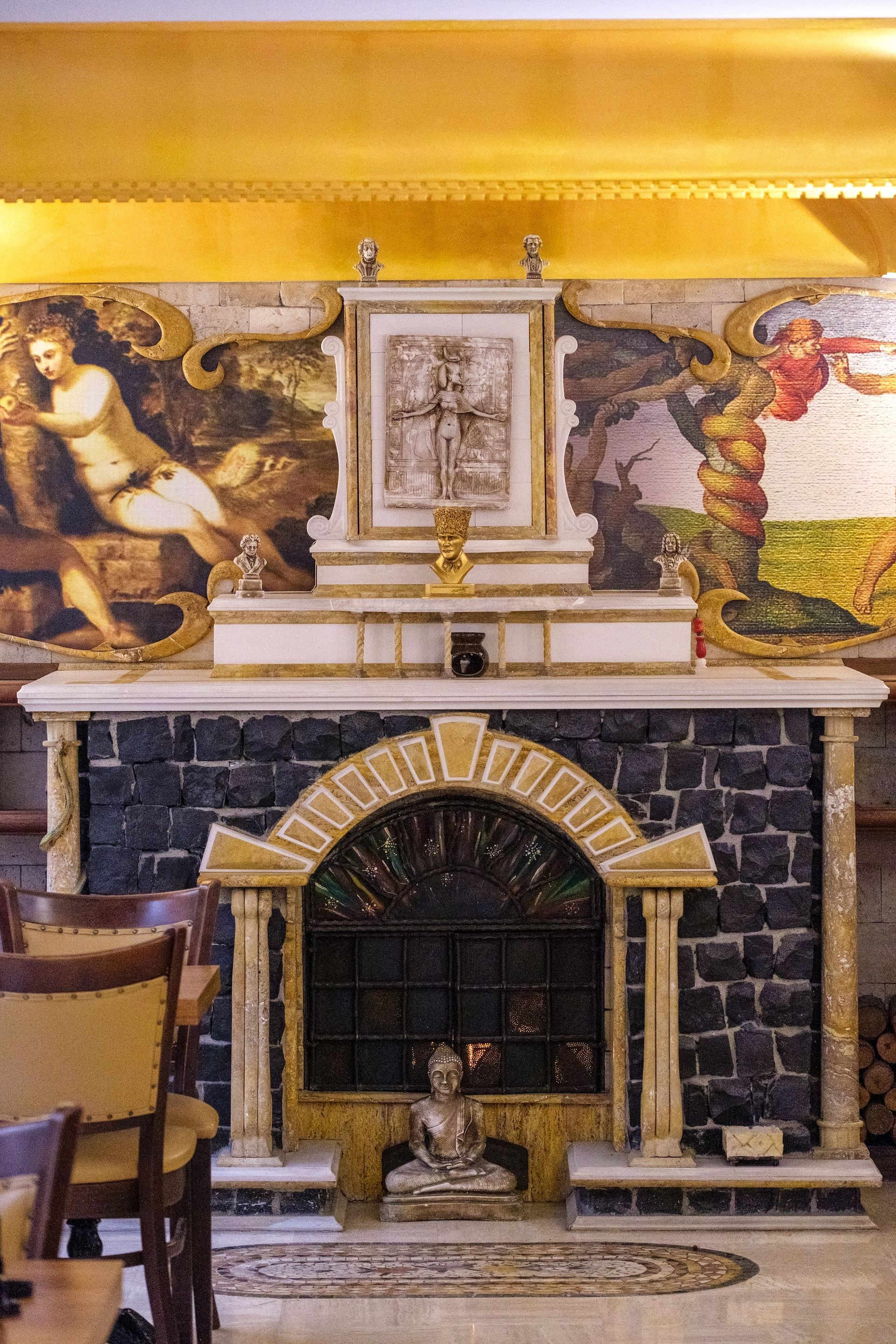 A fireplace with a dark stone hearth, a central gold-framed print, and murals featuring classical figures on the wall.