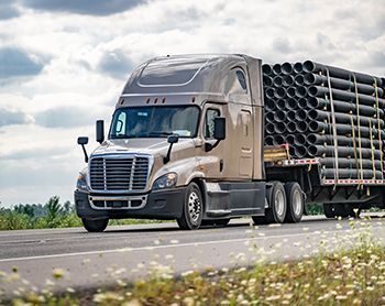 Tan semi-truck hauling a flatbed trailer loaded with black pipes on a highway, under a cloudy sky.