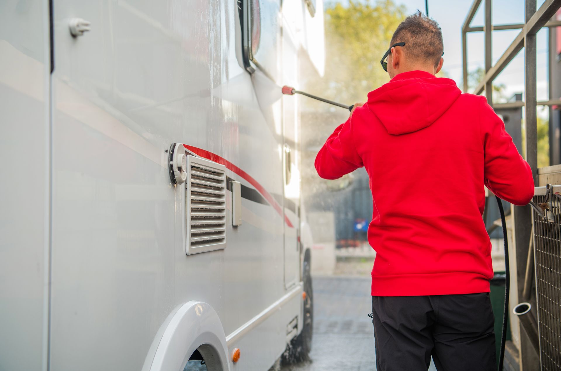 Person in a red hoodie using a pressure washer to clean the side of a white van outdoors