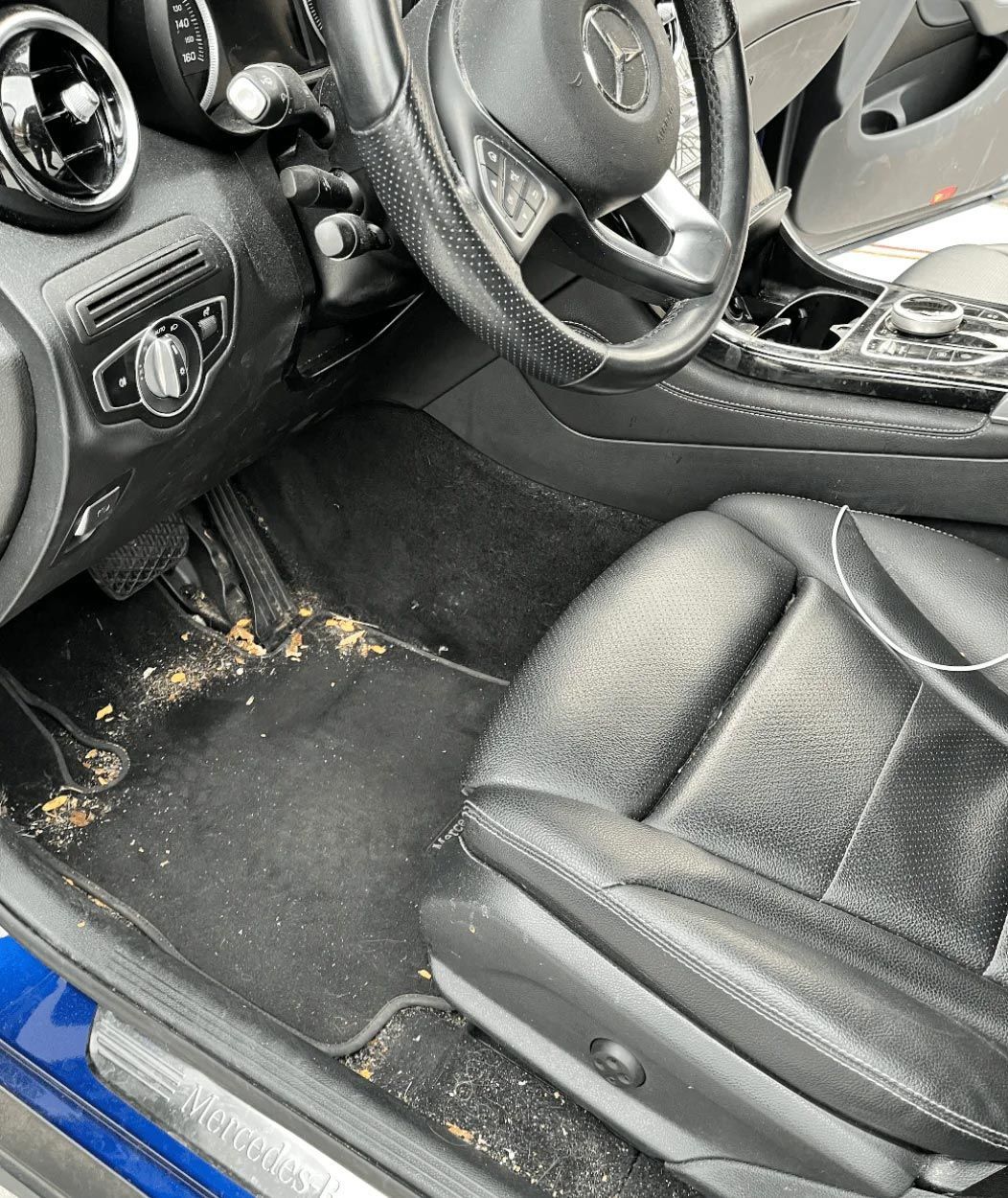 Dirty car interior with debris on the floor mat and black leather driver’s seat.