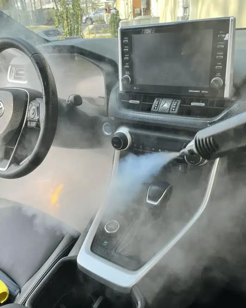 Car interior with steering wheel, dashboard screen, and thick smoke or steam filling the cabin.