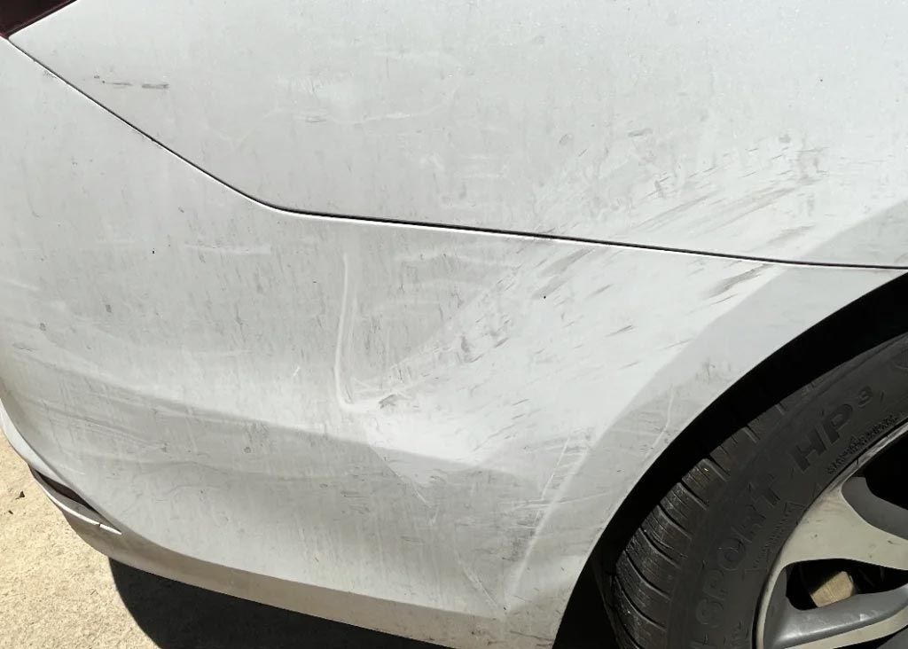 Close-up of a white car’s rear side panel with scuff marks and scratches near the wheel