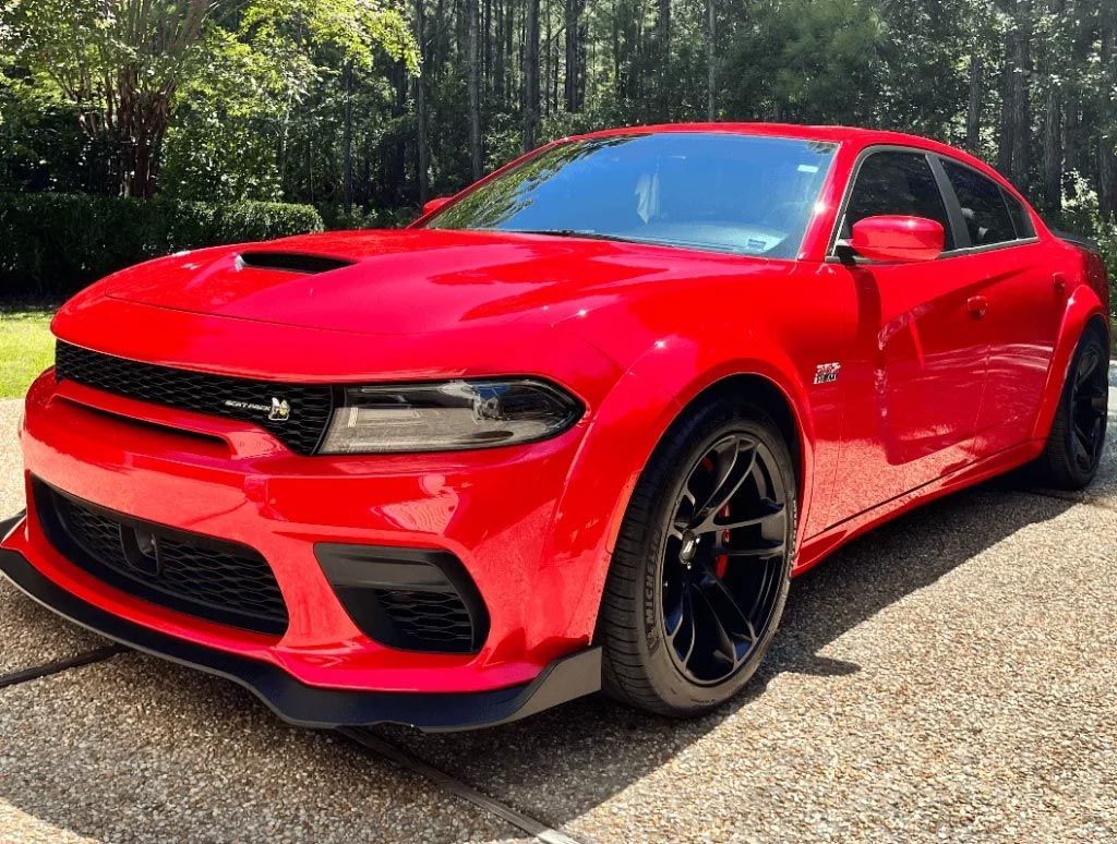 Red Dodge Charger parked on gravel with black rims, viewed from the front in a wooded area