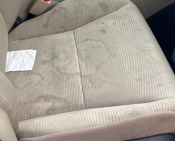 Beige fabric car seat with visible dark stains and a small white tissue on the cushion.